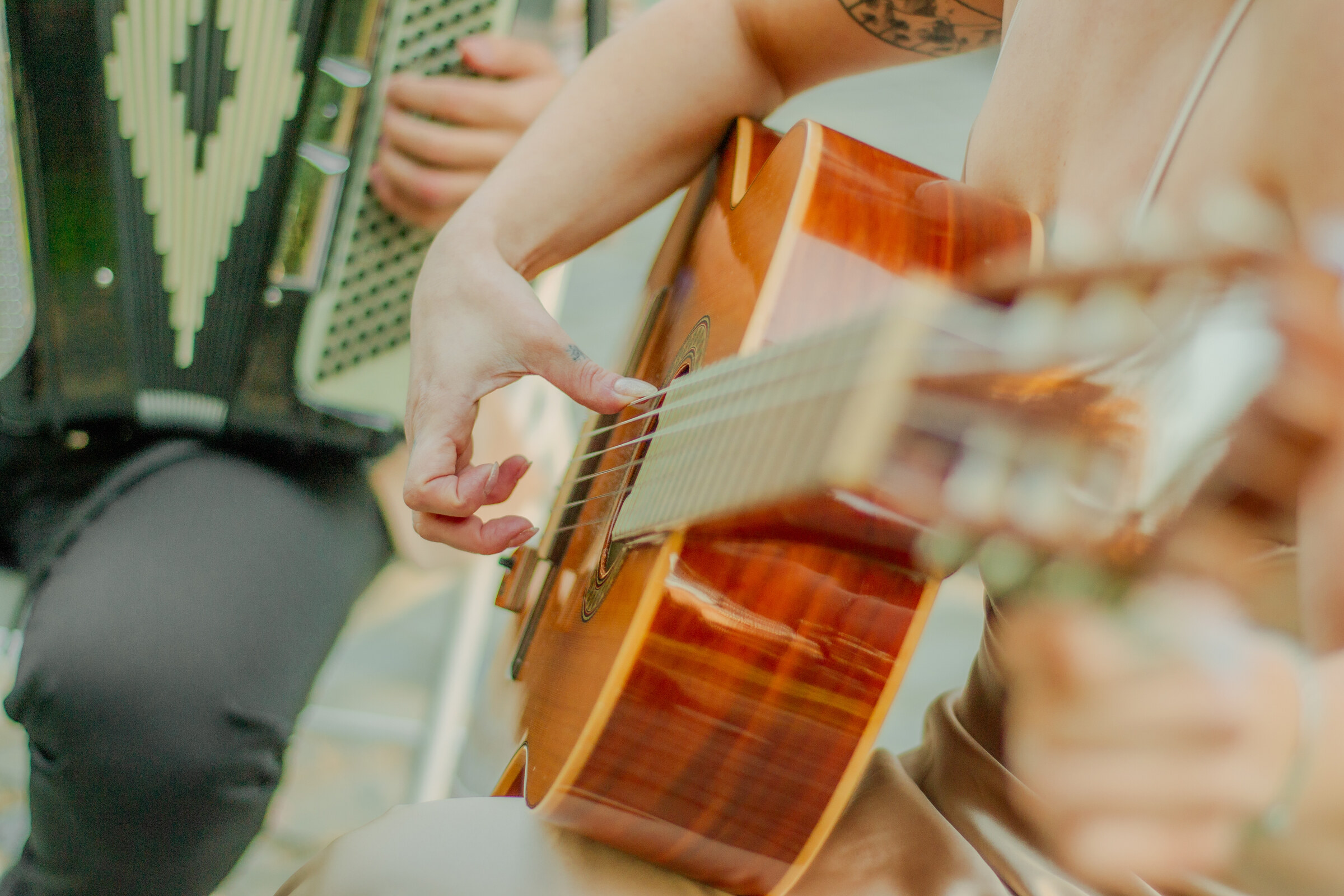Manos femeninas tocando las cuerdas de una guitarra acústica, con un acordeón de fondo desenfocado.